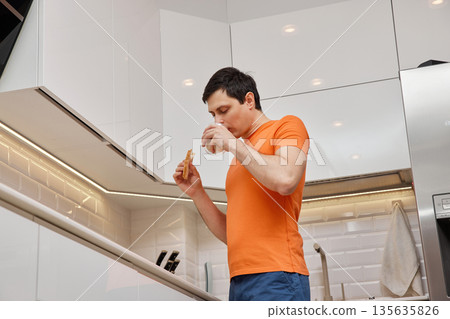 Young man standing in a modern kitchen, eating a sandwich and drinking milk 135635826