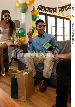 Man opening gift at birthday party, smiling with friends in living room Man opening gift at birthday party, smiling with friends in living room 135635978