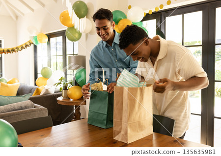 Friends celebrating at home, opening gift bags with excitement and joy 135635981