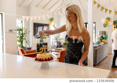 Blonde woman celebrating 21st birthday, cutting cake in decorated living room 135636006