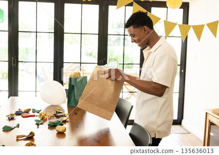 Man preparing gift bags at home for festive celebration, smiling and enjoying Man preparing gift bags at home for festive celebration, smiling and enjoying 135636013