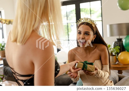 Celebrating birthday, woman giving gift to friend wearing party headband at home 135636027