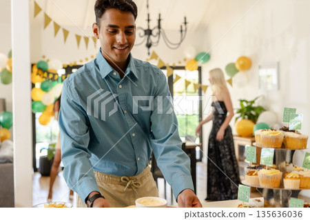 Smiling man in blue shirt arranging desserts at festive birthday party 135636036