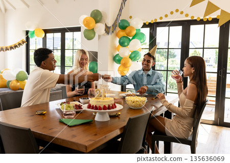 Friends celebrating birthday with cake and drinks at home, smiling and toasting 135636069