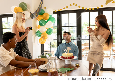 Friends celebrating birthday with cake and balloons, smiling and clapping indoors 135636070