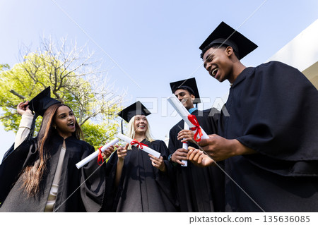 Celebrating success outdoors, graduates in caps and gowns holding diplomas, smiling Celebrating success outdoors, graduates in caps and gowns holding diplomas, smiling 135636085