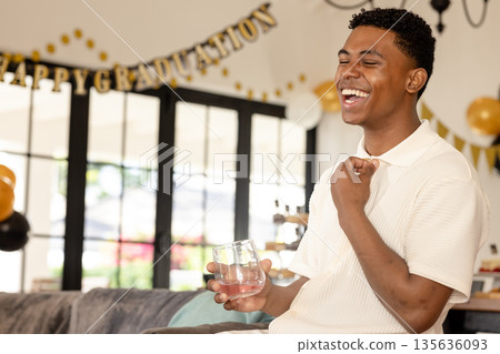 Young man celebrating graduation at home, laughing and holding glass 135636093