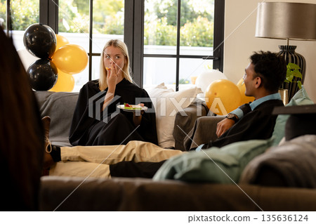 Graduates in gowns relaxing on sofa, enjoying snacks at home celebration Graduates in gowns relaxing on sofa, enjoying snacks at home celebration 135636124