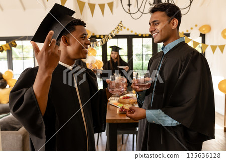 Graduation celebration, two young men in caps and gowns toasting with drinks 135636128
