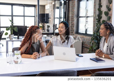 Diverse businesswomen discussing project at office table with laptop and notebooks Diverse businesswomen discussing project at office table with laptop and notebooks 135636201