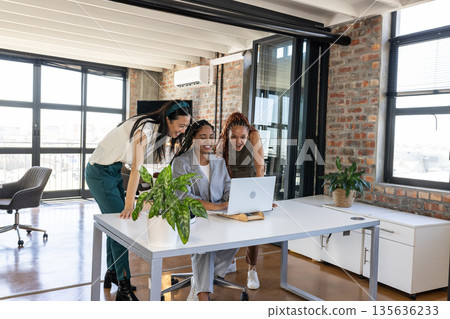 Three women collaborating on laptop in modern office, celebrating successful project 135636233