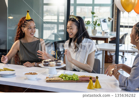 Celebrating in office, colleagues wearing party hats and sharing cupcakes joyfully 135636282