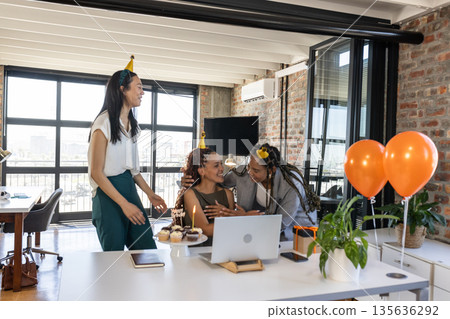 Celebrating birthday in office, colleagues wearing party hats and holding cupcakes 135636292