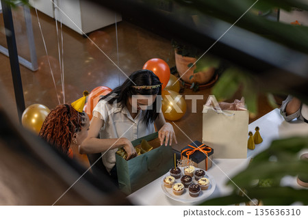Opening gift bag, woman celebrating office party with cupcakes and balloons 135636310