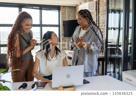 Colleagues celebrating success around laptop in modern office, expressing excitement 135636325