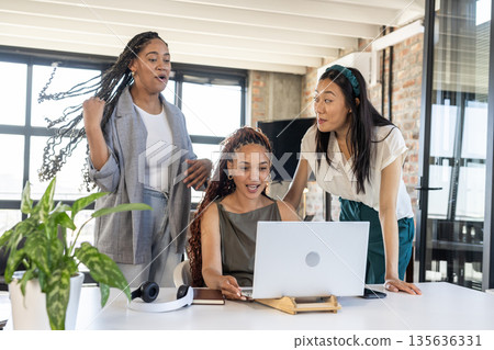 Women collaborating and smiling around laptop, celebrating success in modern office Women collaborating and smiling around laptop, celebrating success in modern office 135636331