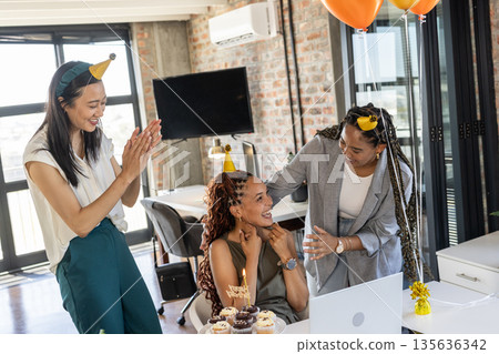 Celebrating birthday in office, colleagues wearing party hats and sharing cupcakes joyfully 135636342