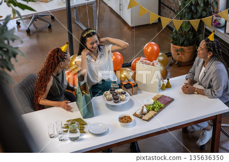 Celebrating at office, diverse women enjoying party with gifts and snacks 135636350