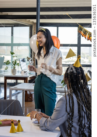 Asian woman holding glass, smiling during office celebration with colleagues 135636351