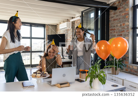 Colleagues celebrating birthday in office with cake, balloons, and party hats 135636352