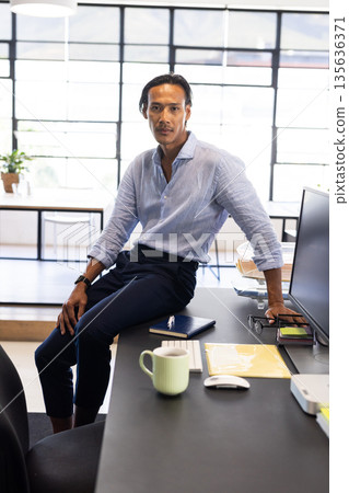 Confident businessman sitting on desk in modern office, looking at camera 135636371