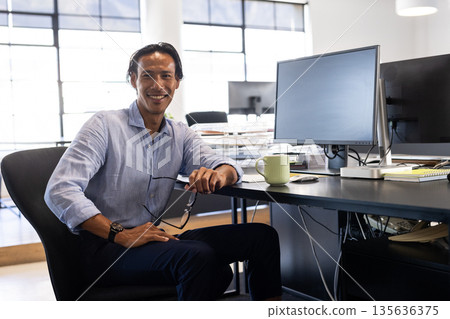 Confident businessman sitting at desk in modern office, holding glasses, smiling 135636375