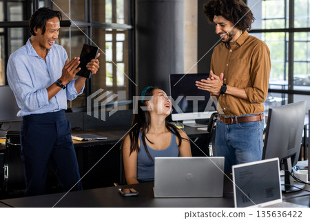 Asian man taking photo of colleagues laughing in modern office workspace 135636422