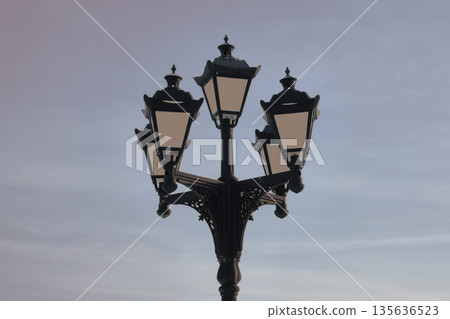 An ornate street lamp with five lanterns is set against a muted sky. The classic design and gentle light create a calming, dreamlike ambiance in an outdoor location An ornate street lamp with five lanterns is set against a muted sky. The classic design and gentle light create a calming, dreamlike ambiance in an outdoor location 135636523