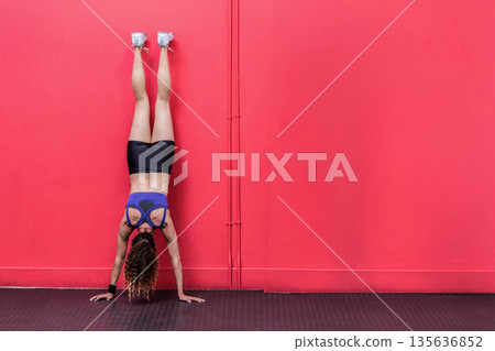 Female fitness enthusiast performing handstand in studio against red wall and wearing wristband 135636852