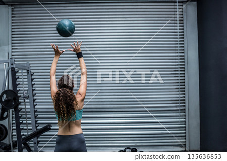 Woman tossing medicine ball against roll-up door in gym garage with weight rack, bench 135636853