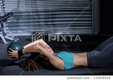 Middle-aged woman performing exercise on black mat in gym holding green medicine ball in teal bra 135636854