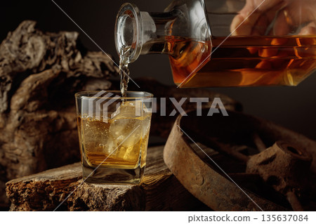Whiskey is poured from a decanter into a glass with ice. 135637084