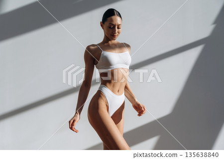 Against the white wall, standing and posing. Beautiful young woman with slim body type is indoors 135638840