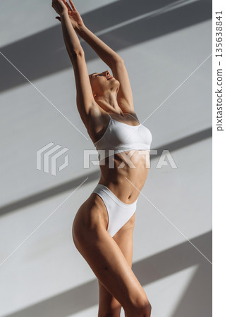 Against the white wall, standing and posing. Beautiful young woman with slim body type is indoors 135638841