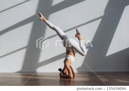 Headstand, doing exercise. Beautiful young woman with slim body type is indoors 135639004