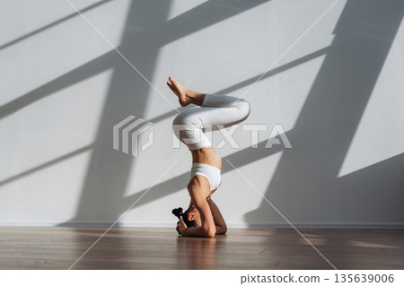 Headstand, doing exercise. Beautiful young woman with slim body type is indoors Headstand, doing exercise. Beautiful young woman with slim body type is indoors 135639006