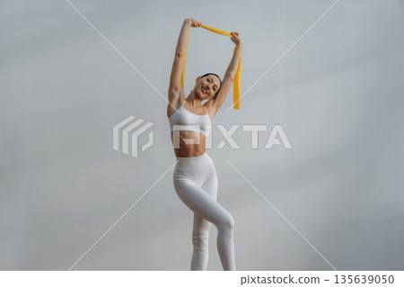 Yellow fitness tape for stretching. Beautiful young woman with slim body type is indoors Yellow fitness tape for stretching. Beautiful young woman with slim body type is indoors 135639050