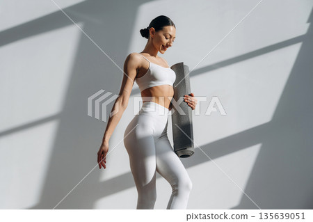 Gray yoga mat in hand. Beautiful young woman with slim body type is indoors Gray yoga mat in hand. Beautiful young woman with slim body type is indoors 135639051