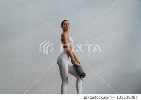 Fitness trainer is holding yoga mat. Beautiful young woman with slim body type is indoors Fitness trainer is holding yoga mat. Beautiful young woman with slim body type is indoors 135639067
