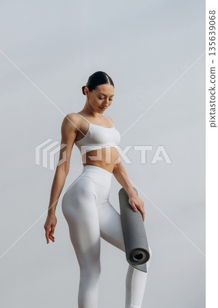 Fitness trainer is holding yoga mat. Beautiful young woman with slim body type is indoors Fitness trainer is holding yoga mat. Beautiful young woman with slim body type is indoors 135639068