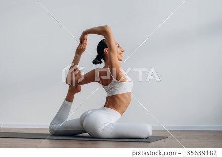 Enjoying doing yoga. Beautiful young woman with slim body type is indoors Enjoying doing yoga. Beautiful young woman with slim body type is indoors 135639182
