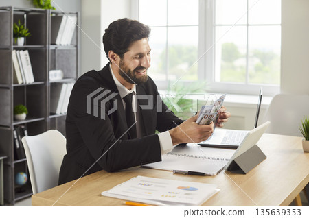 Smiling businessman counting cash by laptop at bright office desk workspace Smiling businessman counting cash by laptop at bright office desk workspace 135639353