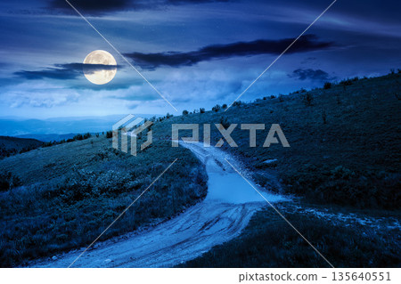 country dirt road through mountains on a summer night. scenic view of rolling hills under dark sky with clouds in full moon light. beautiful alpine meadow for finance investment or abstract concepts 135640551