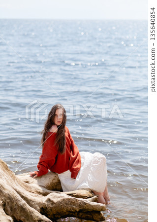 A young woman with long hair and blue eyes, in a white skirt and a terracotta sweater, sits on a tree stump against the backdrop of the sea A young woman with long hair and blue eyes, in a white skirt and a terracotta sweater, sits on a tree stump against the backdrop of the sea 135640854