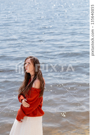 A young woman with long hair and blue eyes, in a white skirt and a terracotta sweater, stands barefoot in the water against the backdrop of the sea A young woman with long hair and blue eyes, in a white skirt and a terracotta sweater, stands barefoot in the water against the backdrop of the sea 135640855