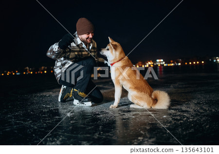 Training the tricks, giving paw. Man with Akita Inu dog is on the ice with skates in winter 135643101