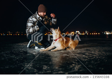 Playing, sitting. Man with Akita Inu dog is on the ice with skates in winter 135643108