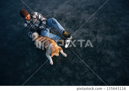 Dark ice, top view. Man with Akita Inu dog is on the ice with skates in winter Dark ice, top view. Man with Akita Inu dog is on the ice with skates in winter 135643116