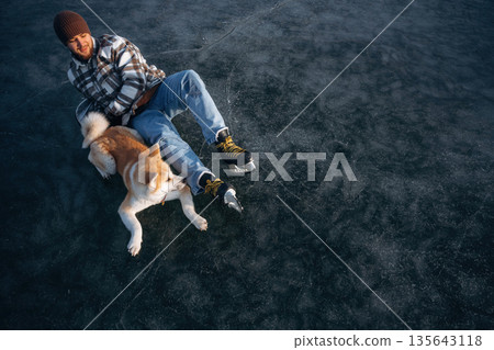 Dark ice, top view. Man with Akita Inu dog is on the ice with skates in winter Dark ice, top view. Man with Akita Inu dog is on the ice with skates in winter 135643118
