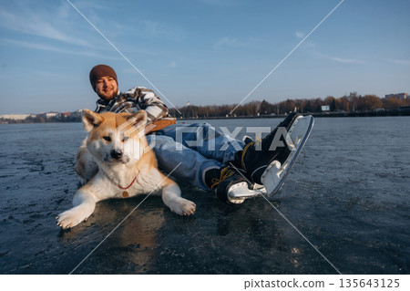 Just lying down and resting. Man with Akita Inu dog is on the ice with skates in winter Just lying down and resting. Man with Akita Inu dog is on the ice with skates in winter 135643125